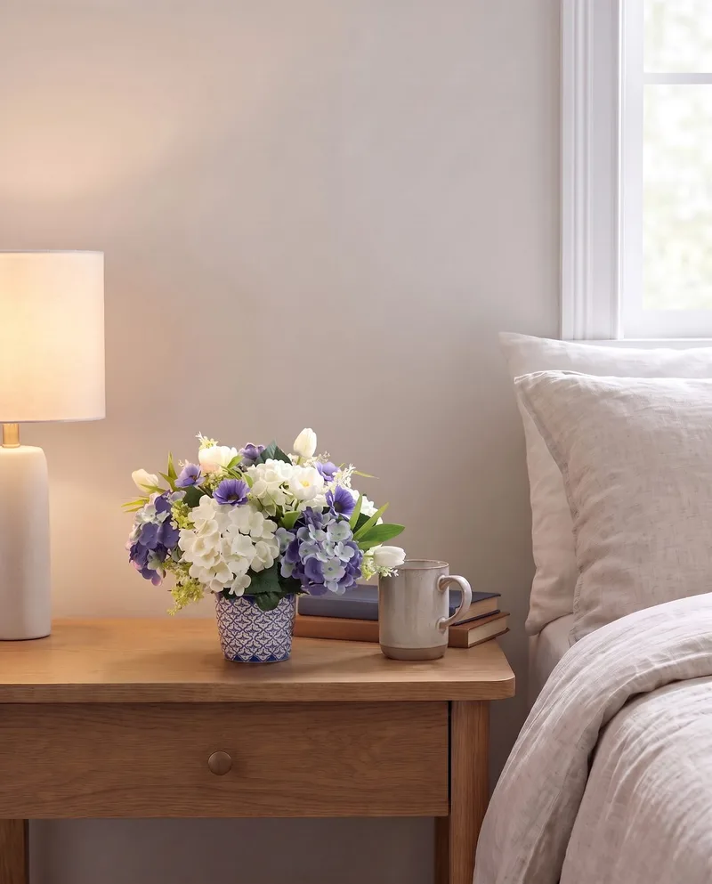 Blissful Blue and White silk flower centerpiece in its Delft pot on a bedroom nightstand beside a lamp and a mug