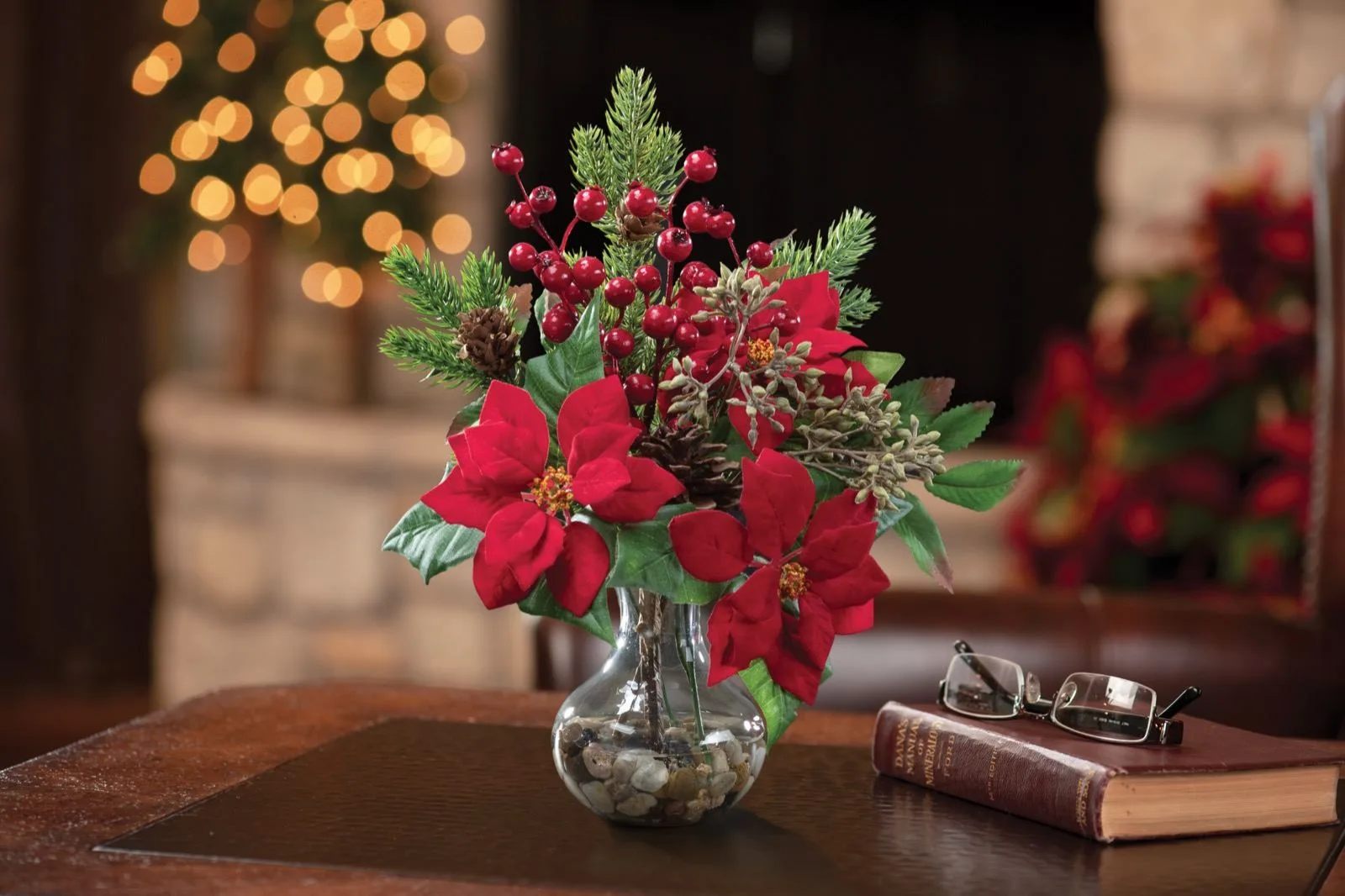 Silk poinsettia, pine and berry Christmas accent on a side table beside a lit tree
