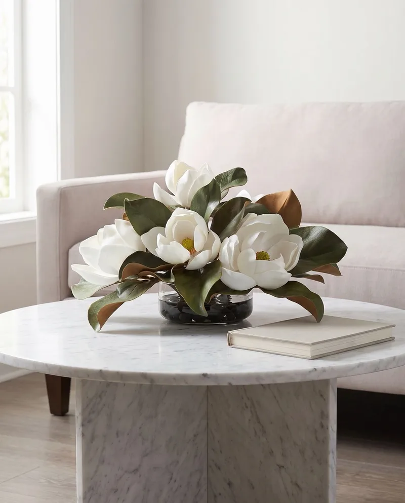 Magnolia silk flower centerpiece on a round marble coffee table beside a hardcover book