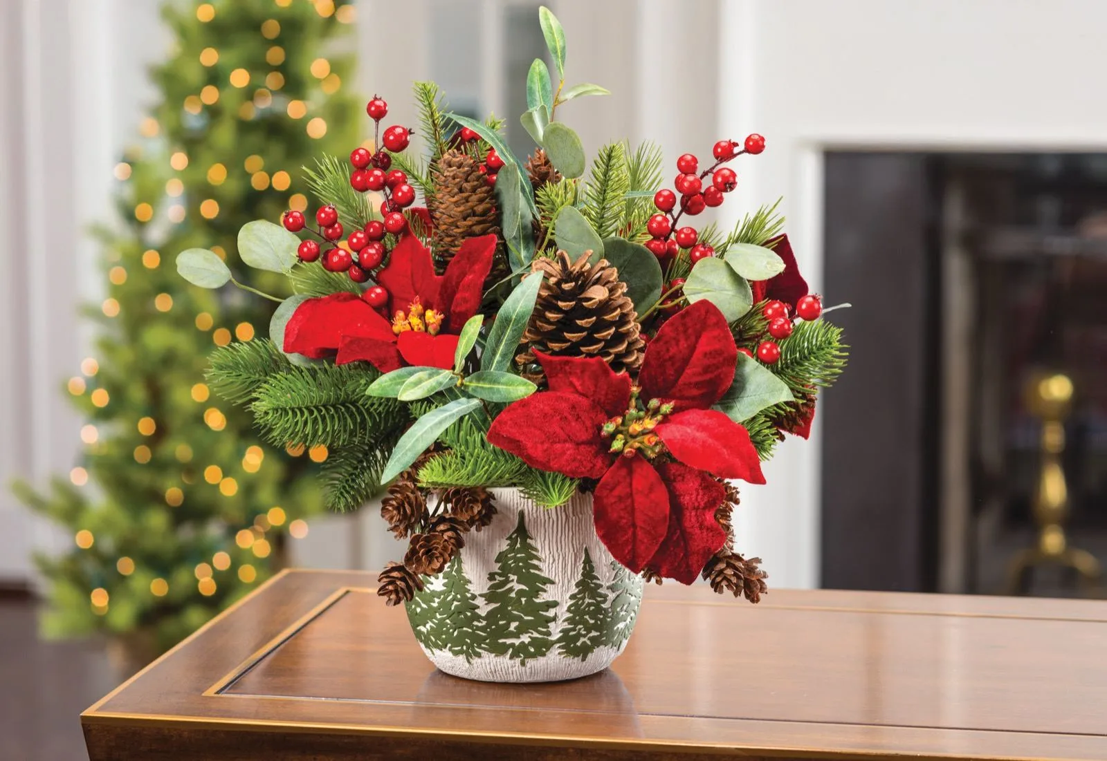Silk velvet poinsettia, pine and berry Christmas bouquet on a wood table beside a lit tree