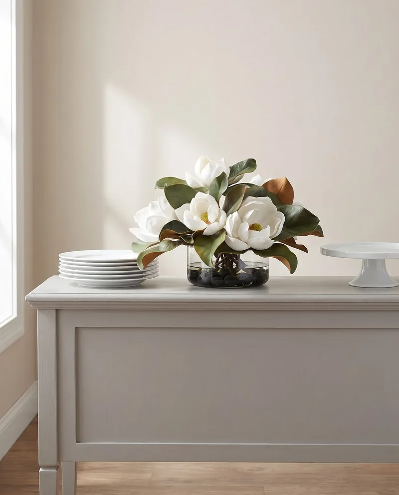 Magnolia silk flower centerpiece on a painted sideboard beside a stack of white plates and a cake stand