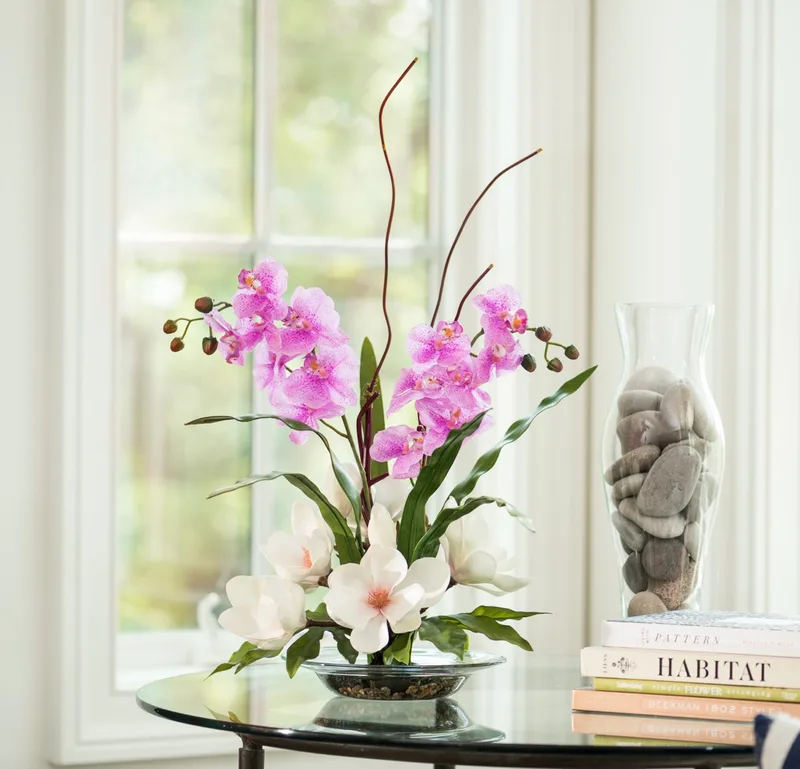 Magnolias and orchids silk flower arrangement on a round glass table beside a window
